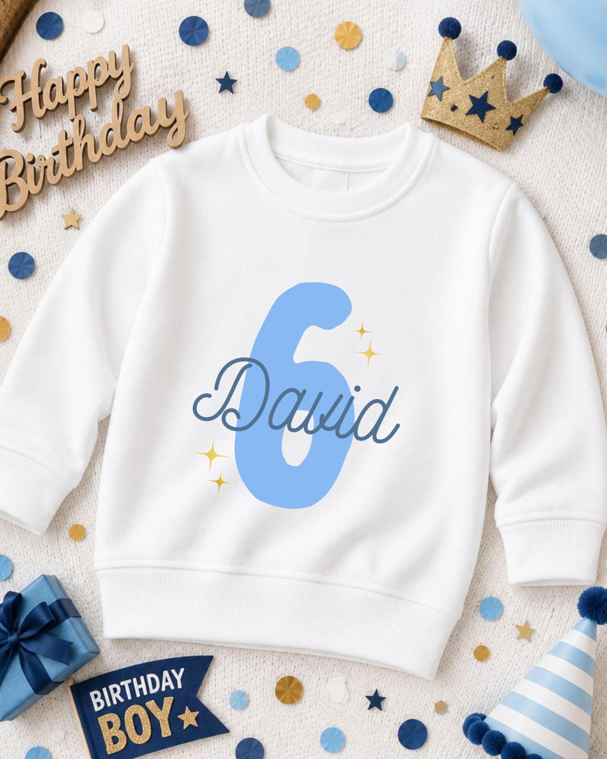 A white sweatshirt with the number 6 and the name "David" printed on it, surrounded by birthday decorations such as a crown, confetti, and a birthday hat.