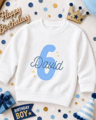 A white sweatshirt with the number 6 and the name "David" printed on it, surrounded by birthday decorations such as a crown, confetti, and a birthday hat.