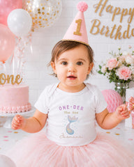 A young girl wearing a pink party hat and a white t-shirt with a mermaid design is posing in front of a birthday cake and balloons.