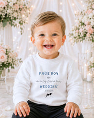 A smiling baby boy wearing a white sweater with text and a graphic design, sitting in front of a floral background.