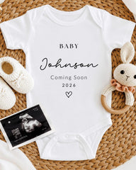 A white baby onesie with the text "BABY Johnson Coming Soon 2026" printed on it, along with a crocheted bunny toy and a black and white photograph of an Ultrasound image.