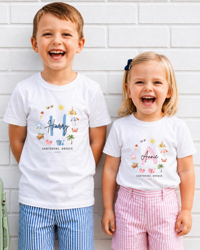 Two children, a boy and a girl, are standing next to each other and smiling. They are wearing matching white t-shirts with colorful illustrations and text.