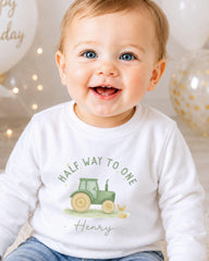 A smiling baby boy wearing a white shirt with a tractor graphic and the text "Half Way to One Henry" printed on it.