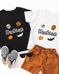 A black t-shirt with the name "Hudson" printed on it, a white t-shirt with the same name, a pair of brown shorts, a pair of black and white checkered shoes, and a pair of sunglasses.