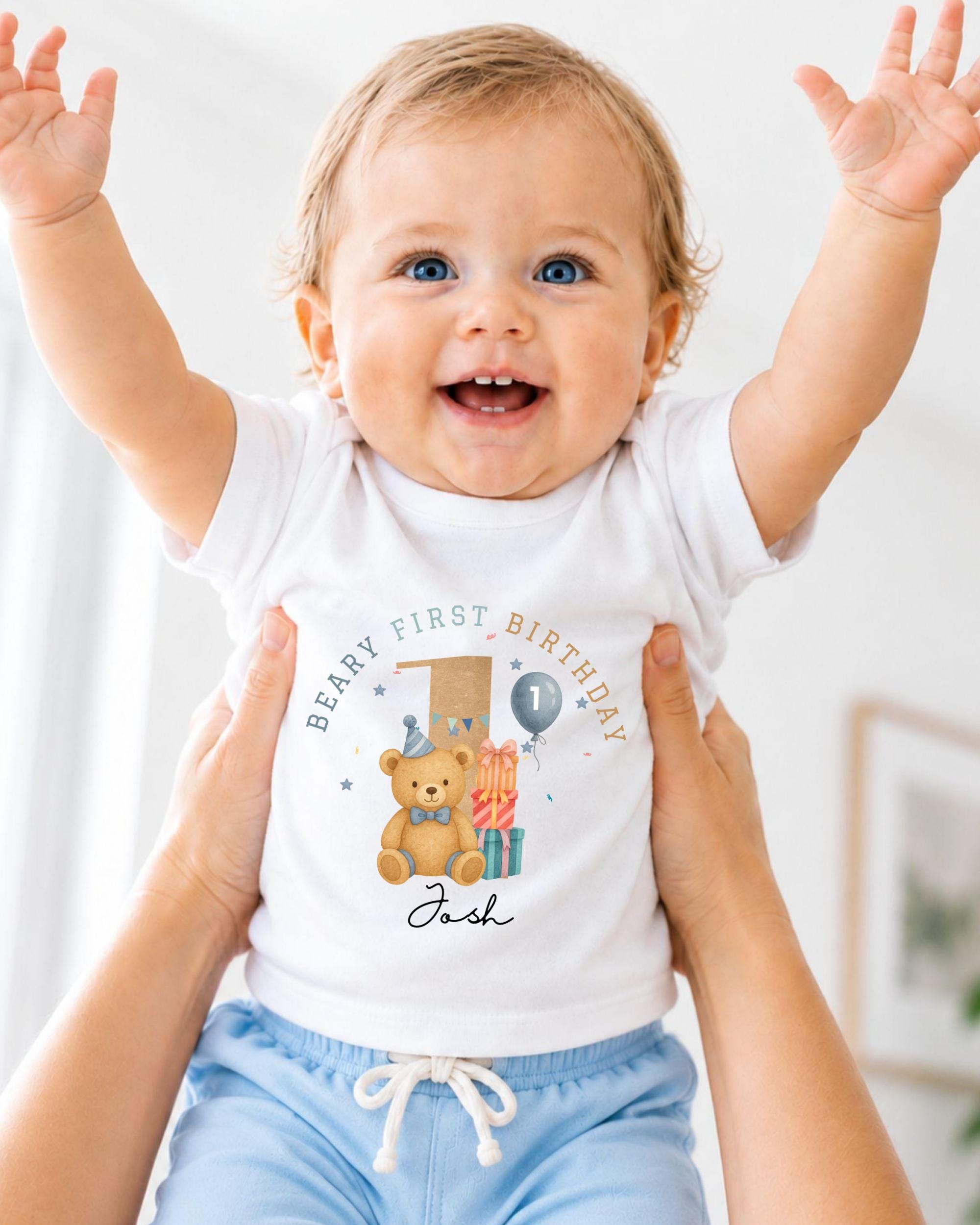 A smiling baby wearing a white t-shirt with a teddy bear and the text "Beary First Birthday" is being held up by two people.