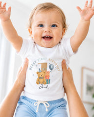 A smiling baby wearing a white t-shirt with a teddy bear and the text "Beary First Birthday" is being held up by two people.