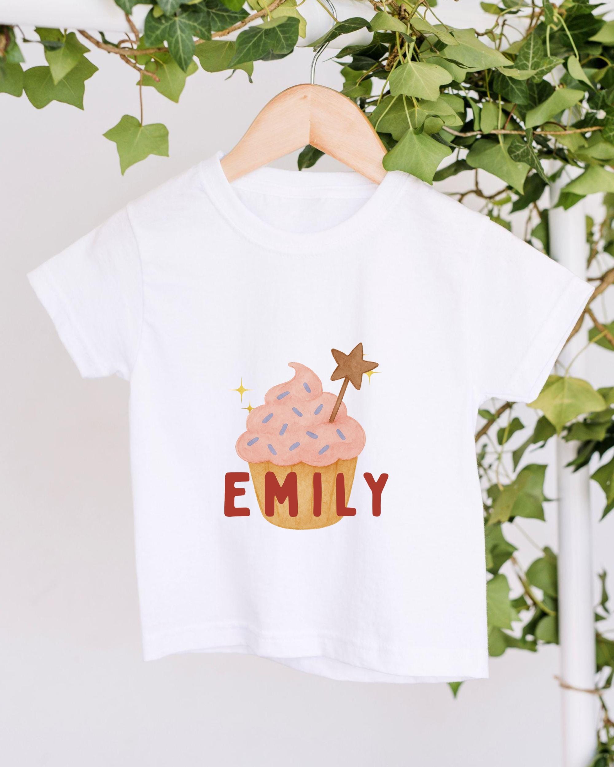 A white t-shirt with a cupcake design and the name "EMILY" printed on it, hanging on a wooden hanger.