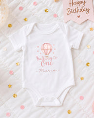 A white onesie with a hot air balloon design and the text "Halfway to One - Maria" printed on it, along with confetti and a birthday card.