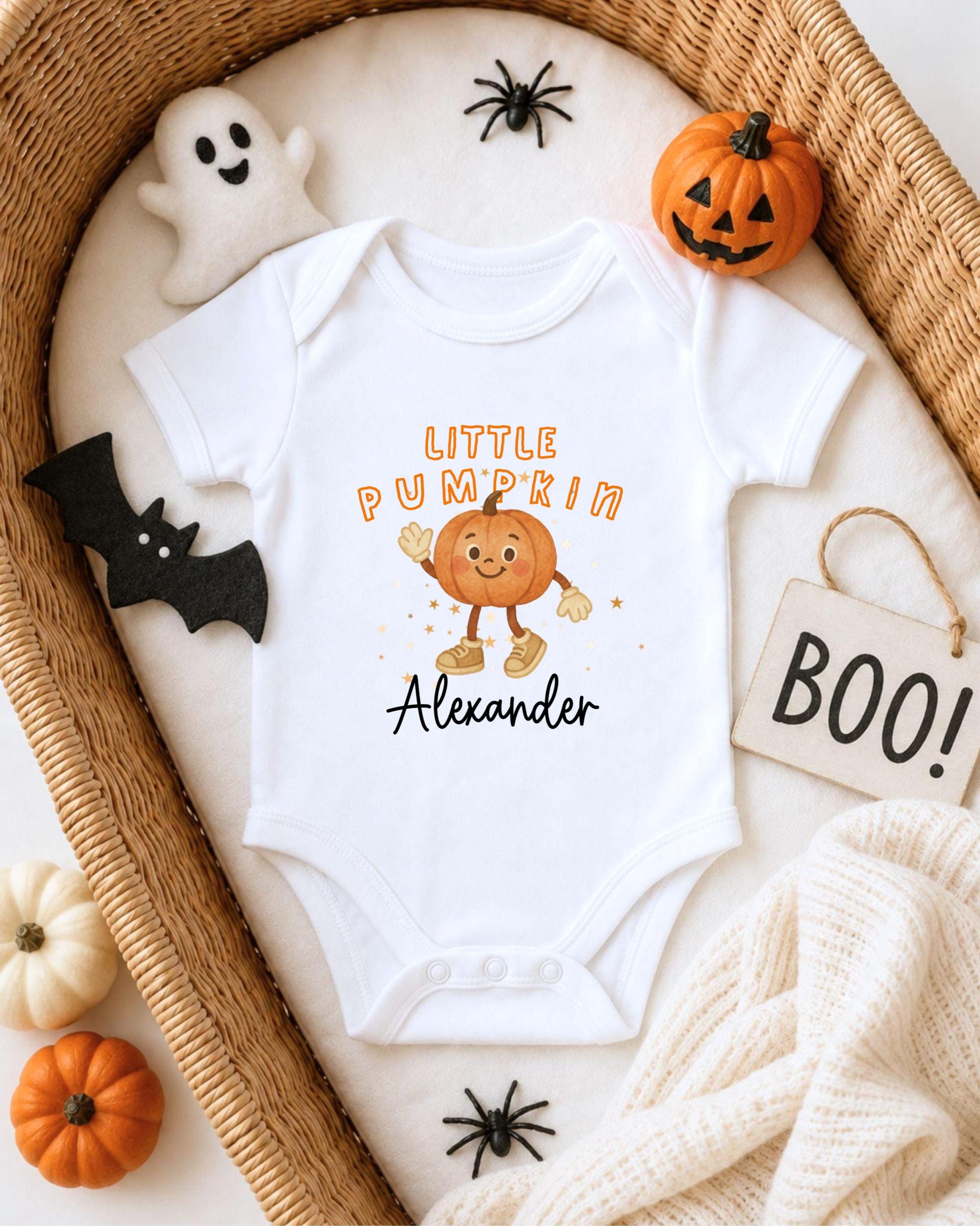 A white onesie with a pumpkin character and the name "Alexander" printed on it, surrounded by Halloween decorations such as a ghost, a pumpkin, and a "Boo!" sign.