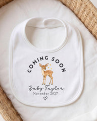 A white baby bib with a printed design of a deer and the text "Coming Soon Baby Taylor November 2027" printed on it.