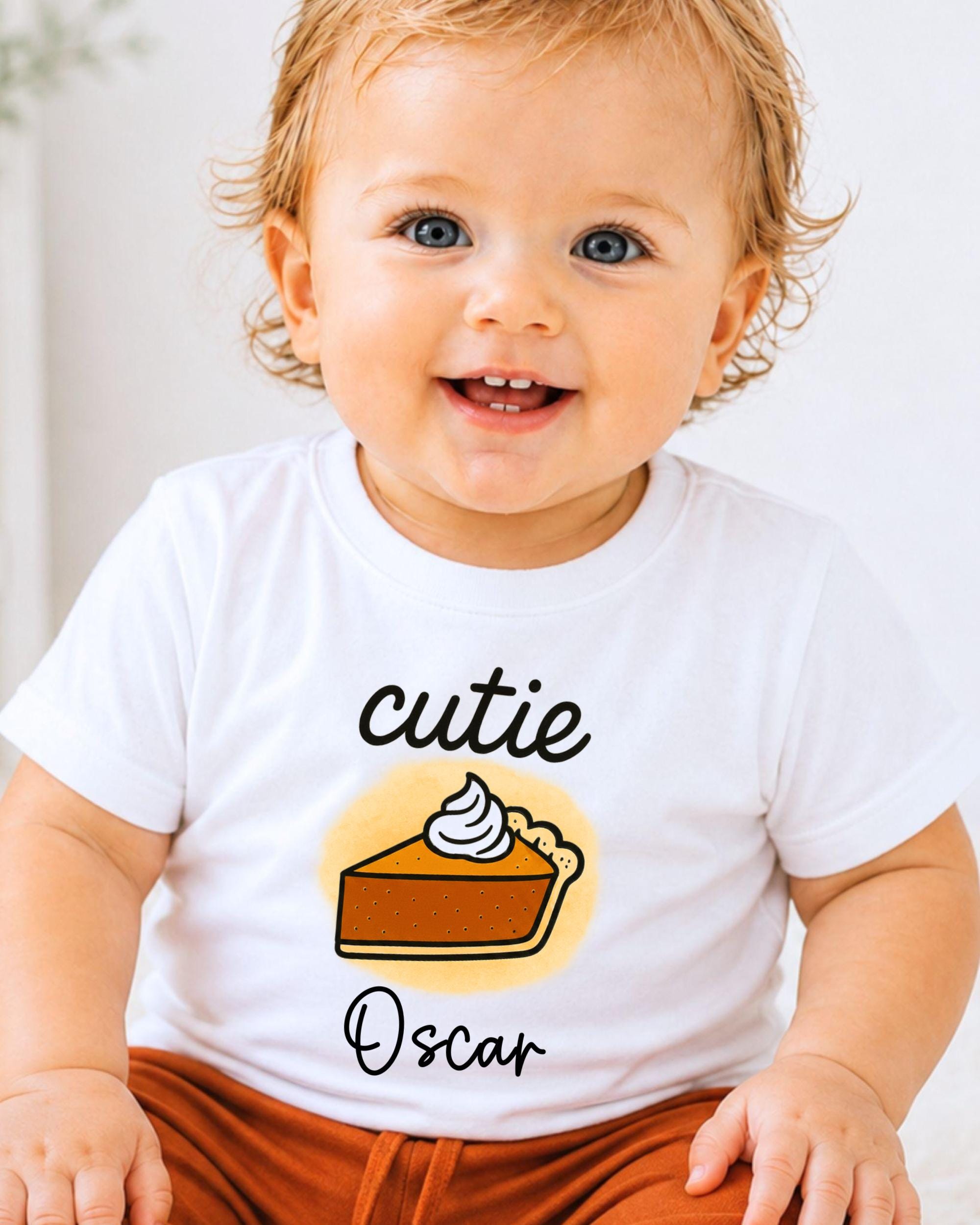 A smiling baby boy wearing a white t-shirt with a pumpkin pie graphic and the name "Oscar" printed on it.