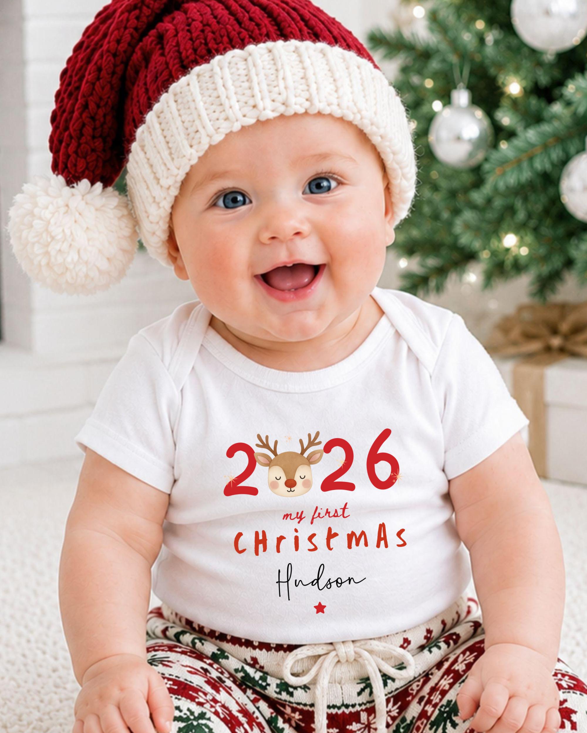 A smiling baby wearing a red and white knit hat and a white t-shirt with the text "26 my first christmas Hudson" is sitting in front of a Christmas tree.