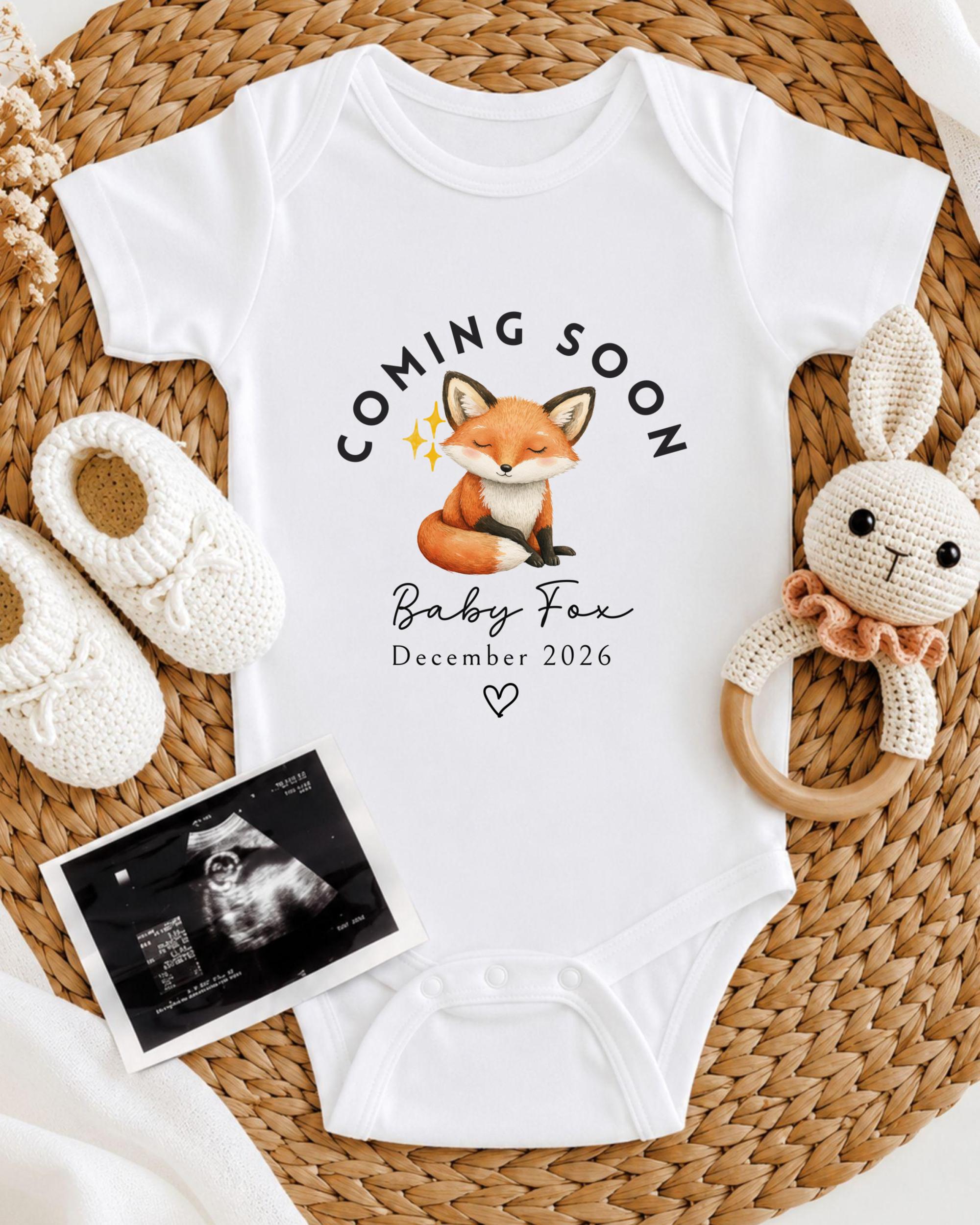A baby announcement outfit with a fox graphic, a crocheted rabbit, and a baby announcement photo, all arranged on a woven placemat.