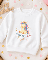A white sweatshirt with a unicorn graphic and the text "Birthday Girl Sophia" printed on it, surrounded by confetti.
