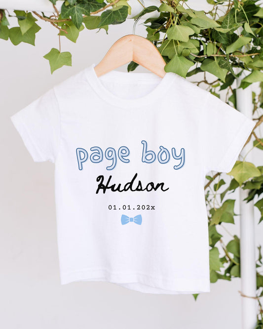 A white t-shirt with the text "Page boy Hudson" printed on it, along with a date "01.01.2022" below it. The t-shirt is hanging on a wooden hanger, and there are green leaves visible in the background.
