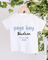 A white t-shirt with the text "Page boy Hudson" printed on it, along with a date "01.01.2022" below it. The t-shirt is hanging on a wooden hanger, and there are green leaves visible in the background.