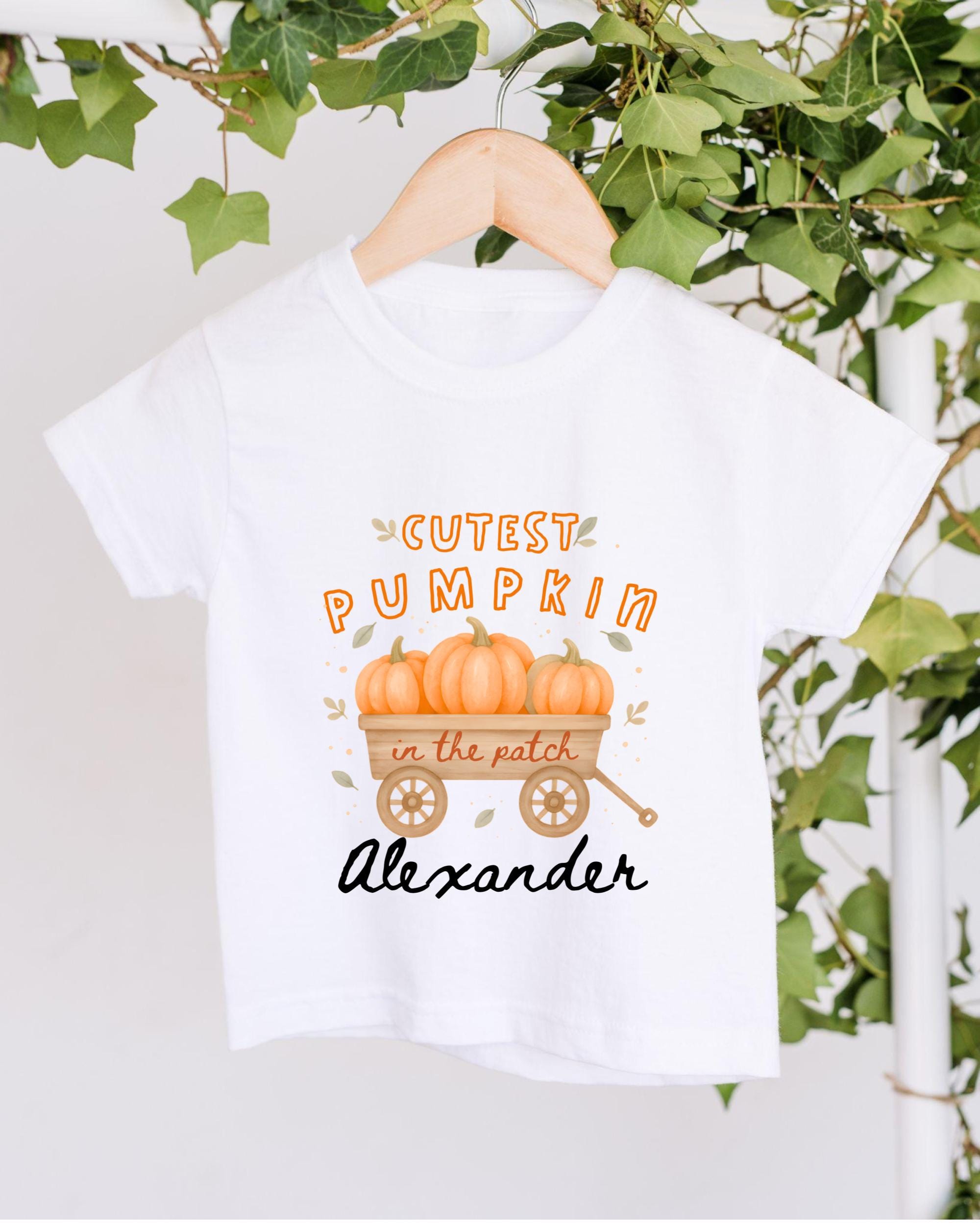 A white t-shirt with a graphic design featuring a wagon filled with pumpkins and the text "Cutest Pumpkin in the Patch Alexander" printed on it.