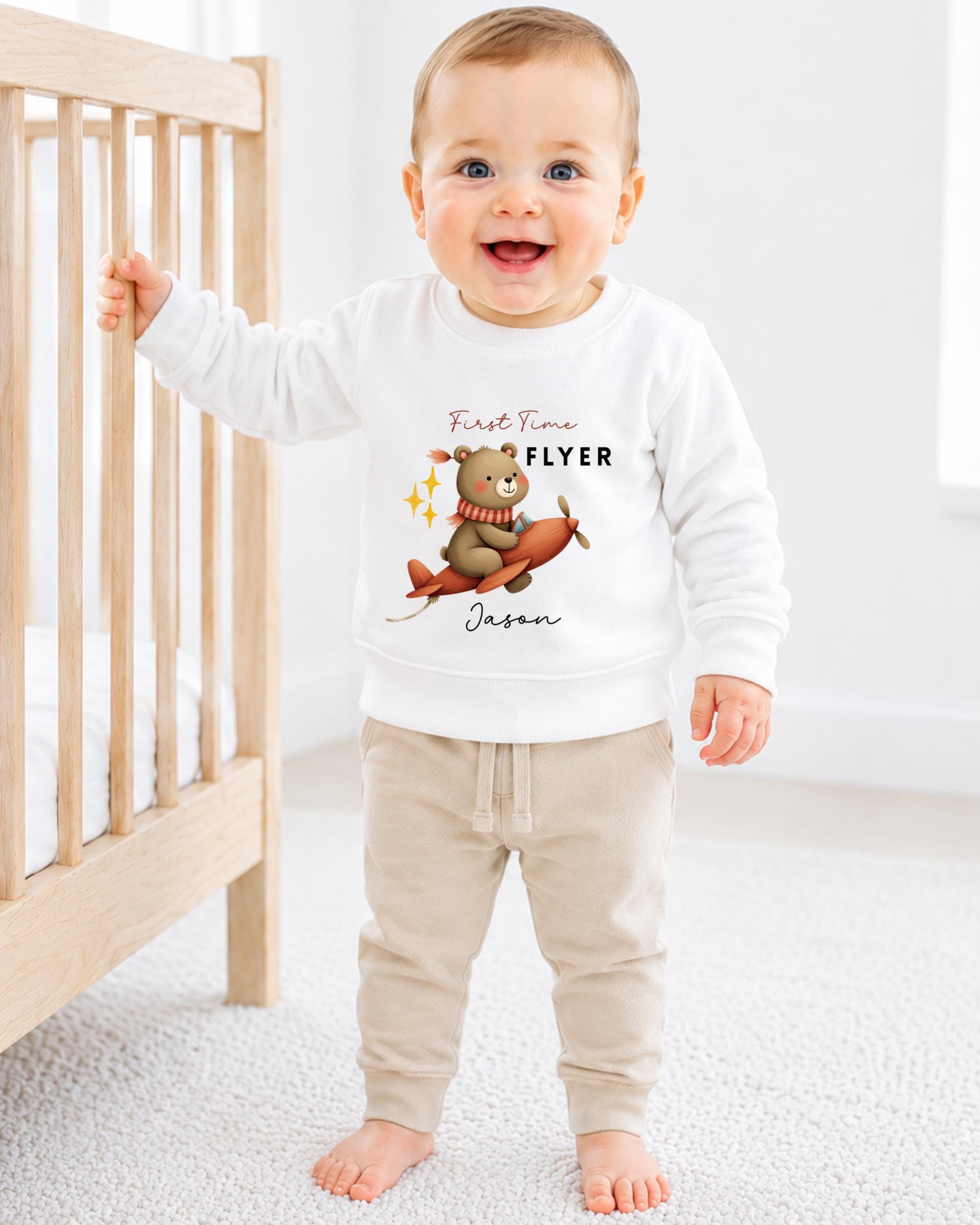 A smiling baby boy wearing a white sweatshirt with a bear graphic and the text "First Time Flyer Jason" is standing in a nursery.