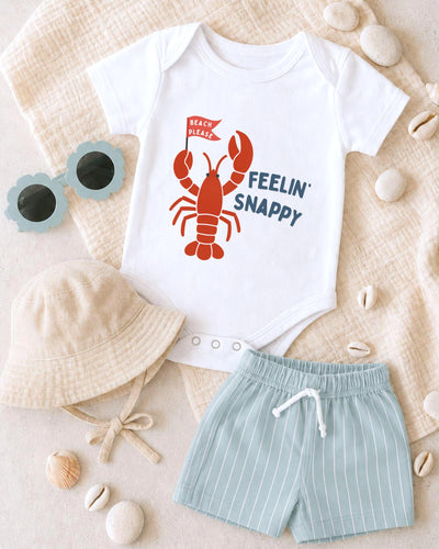 A baby's outfit consisting of a white onesie with a lobster design, a beige bucket hat, and light blue shorts, along with sunglasses and seashells, arranged on a beige surface.