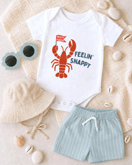 A baby's outfit consisting of a white onesie with a lobster design, a beige bucket hat, and light blue shorts, along with sunglasses and seashells, arranged on a beige surface.