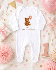 A white onesie with a teddy bear and the text "JANE'S FIRST BIRTHDAY" printed on it, surrounded by pink and gold confetti, a pink balloon, and other baby-related items.