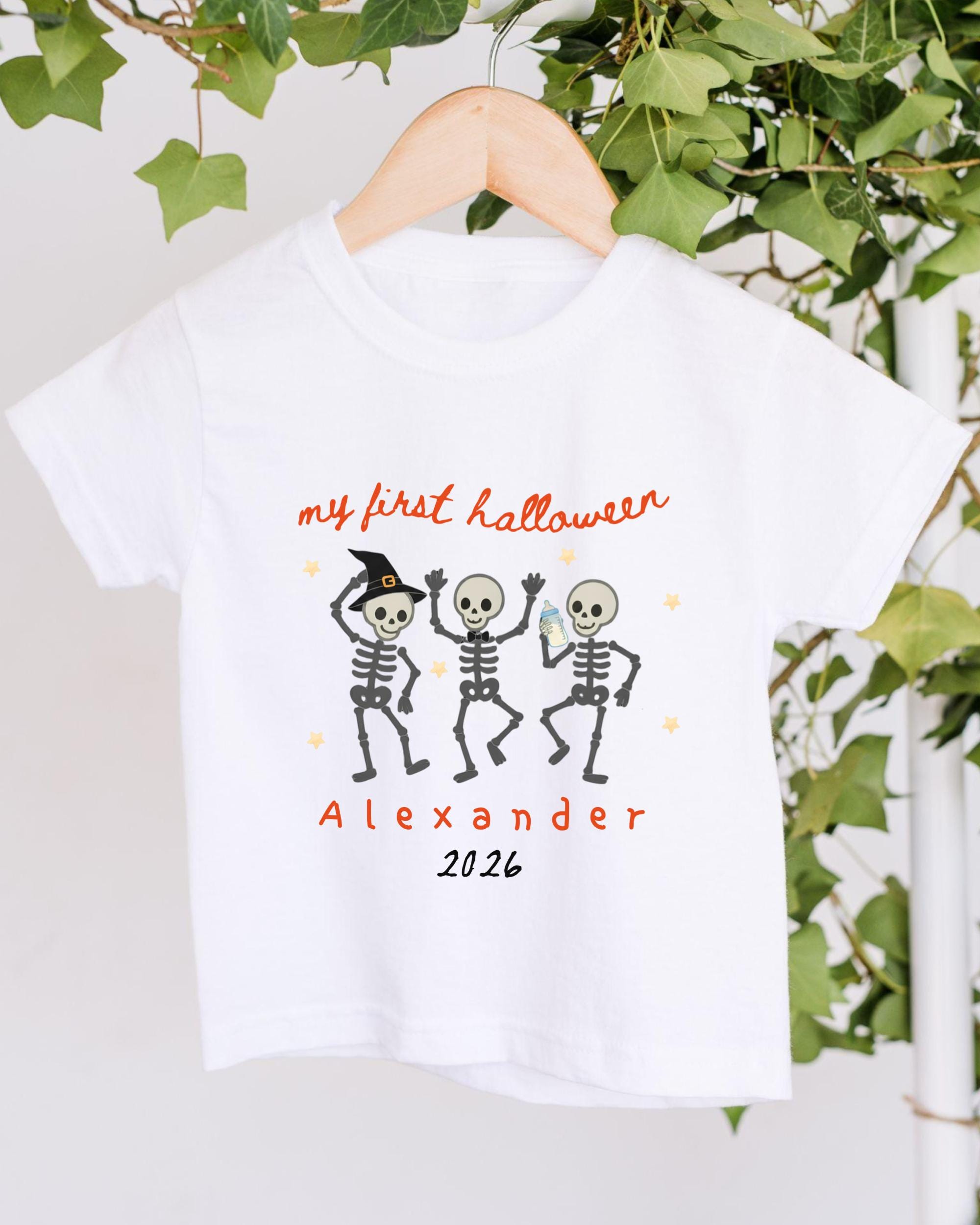 A white t-shirt with a Halloween-themed design featuring three dancing skeletons and the text "my first Halloween Alexander 2026".