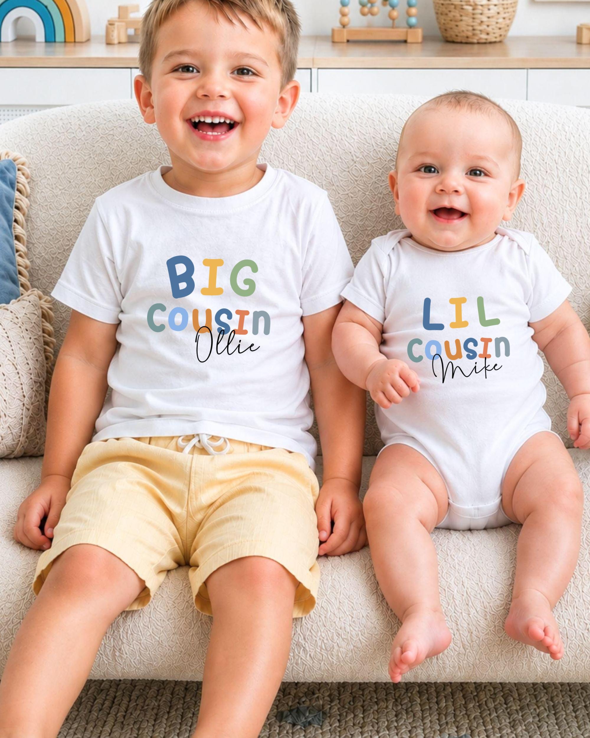 Two young children, a baby and a toddler, sitting on a couch and smiling. They are wearing matching white t-shirts with the text "BIG COUSIN OLLIE" printed on them.