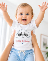 A smiling baby wearing a white t-shirt with a graphic design and text, being held up by someone's arms.