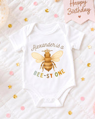 A white baby onesie with a bee design and the text "Alexander is a BEE-SY ONE" printed on it, along with confetti and a birthday card.