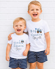 Two young boys wearing matching white t-shirts with blue text and graphics, smiling and posing together against a white brick wall.