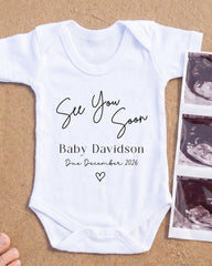 A white baby onesie with text that reads "See You Soon Baby Davidson Due December 2026" printed on it, along with a heart symbol. The onesie is placed next to a photograph of an ultrasound image.