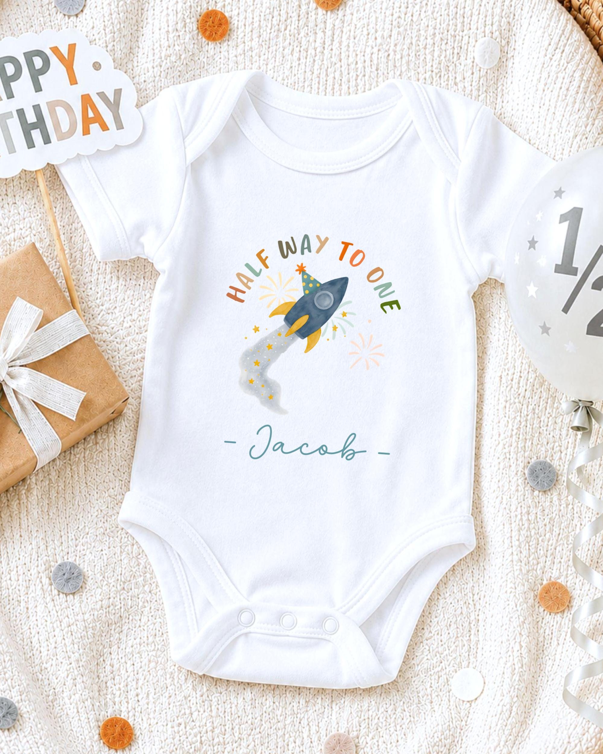 A white onesie with a rocket ship design and the text "Half Way to One" printed on it, along with the name " Jacob" written below. The onesie is placed on a white blanket, surrounded by various party decorations such as a birthday sign, a balloon, and confetti.