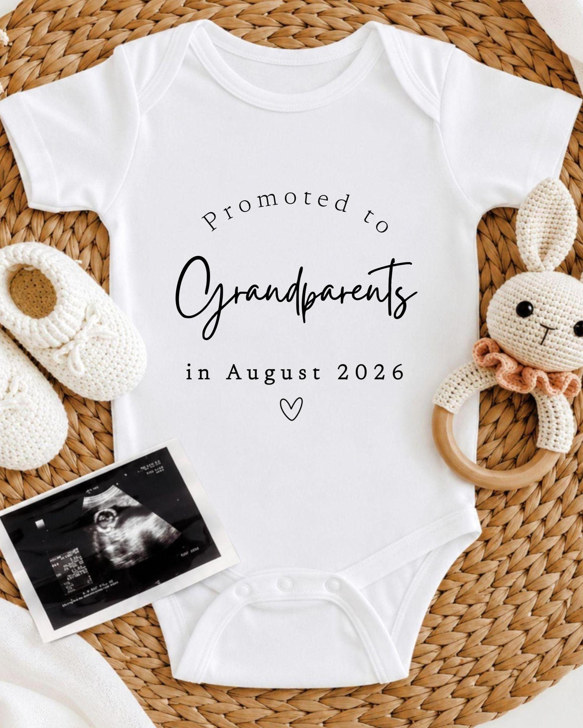 A white baby onesie with the text "Promoted to Grandparents in August 2026" printed on it, along with a crocheted rabbit toy and a black and white photograph of an Ultrasound image.