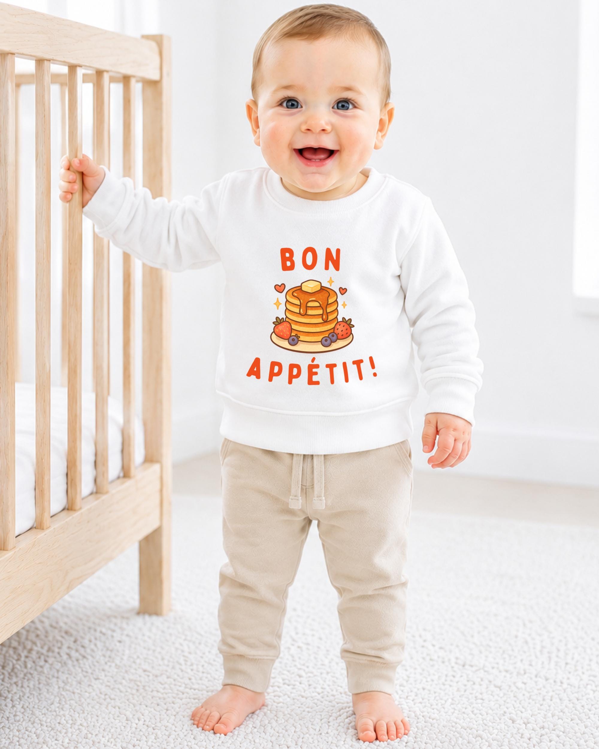 A smiling baby boy wearing a white sweatshirt with a pancake and breakfast design, standing next to a wooden crib.