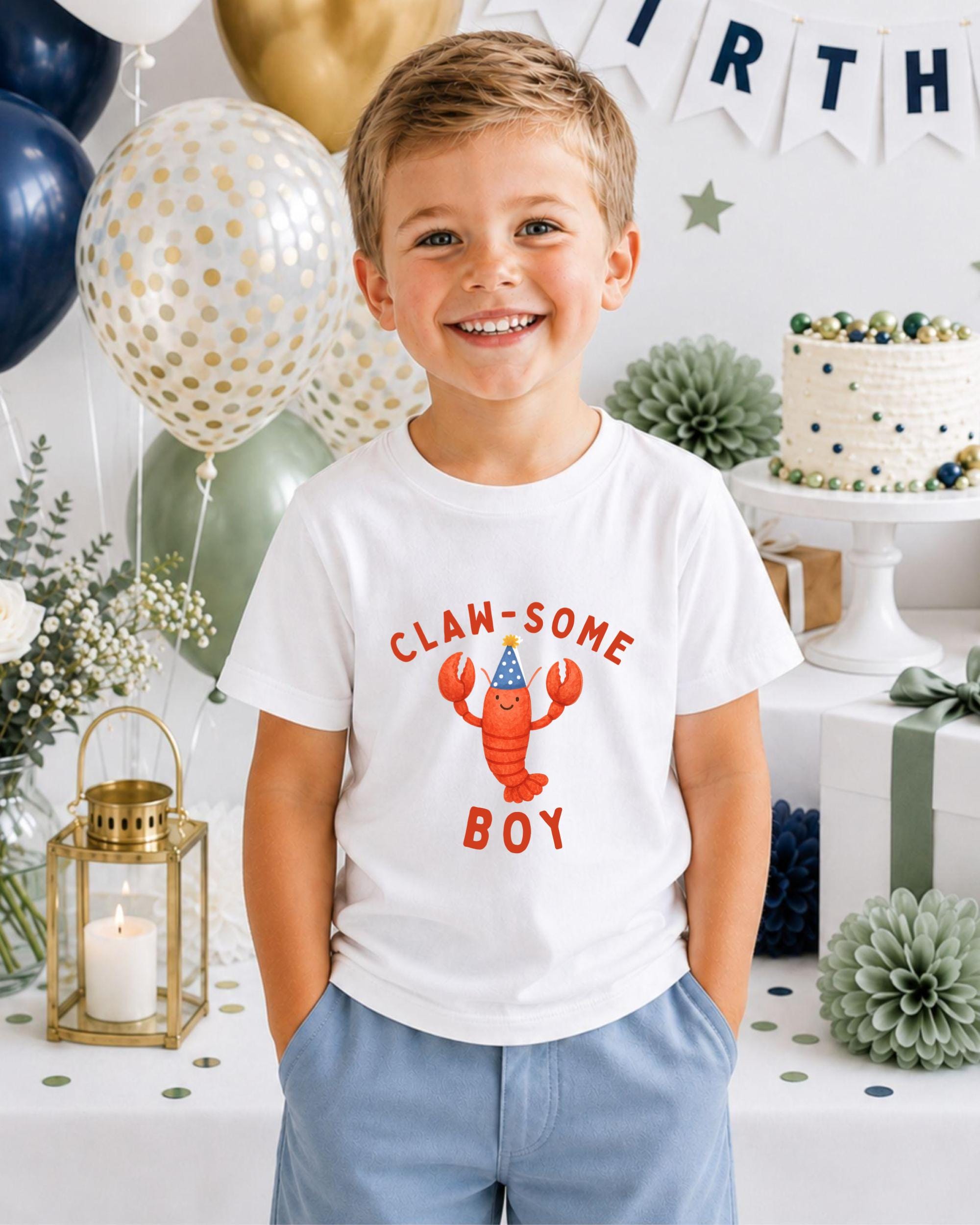 A young boy wearing a white t-shirt with a lobster graphic and the text "CLAW SOME BOY" is smiling at the camera. The background includes balloons, a cake, and other party decorations.