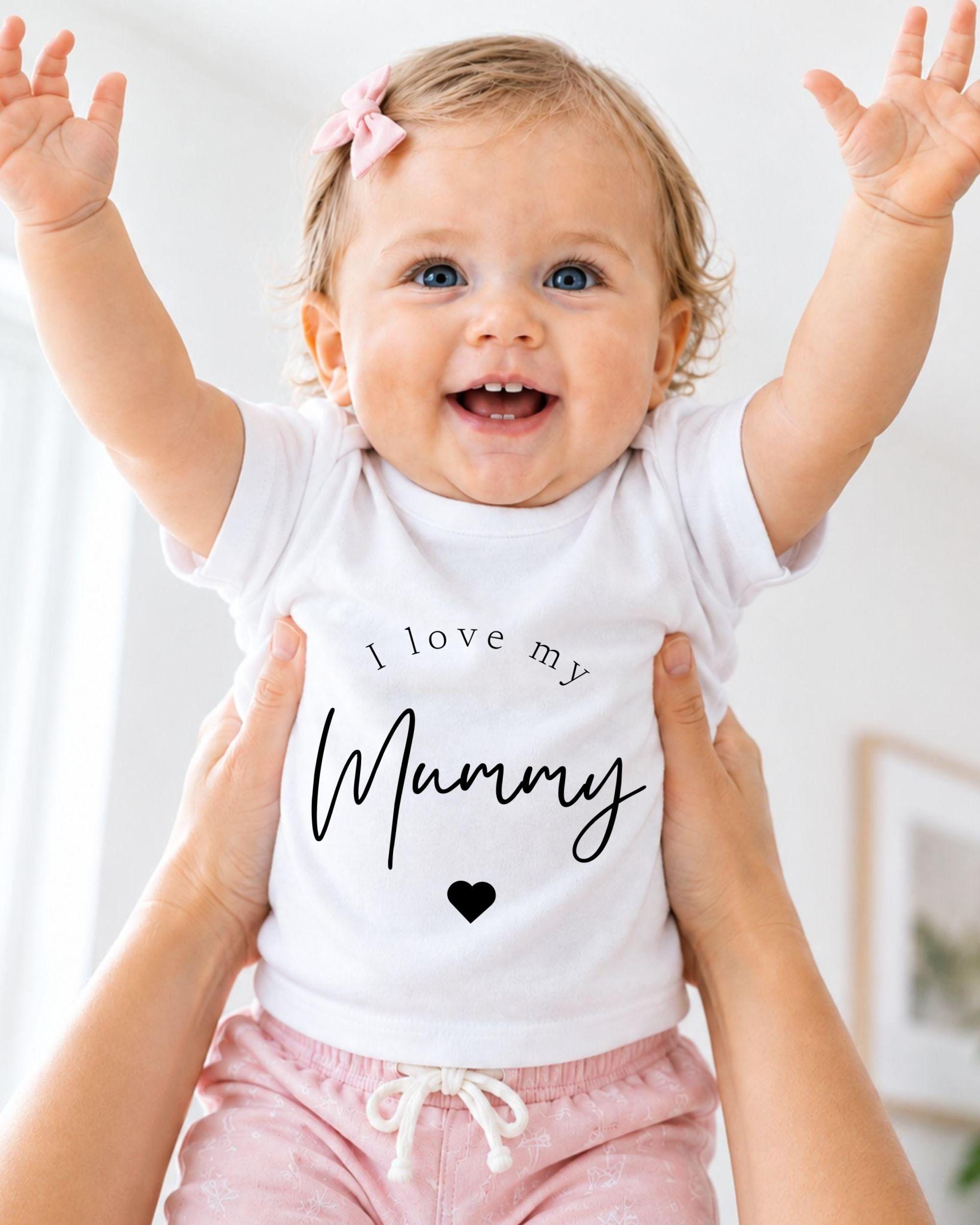 A smiling baby girl wearing a white shirt with the text "I love my Mummy" and pink shorts, being held up by someone's arms.