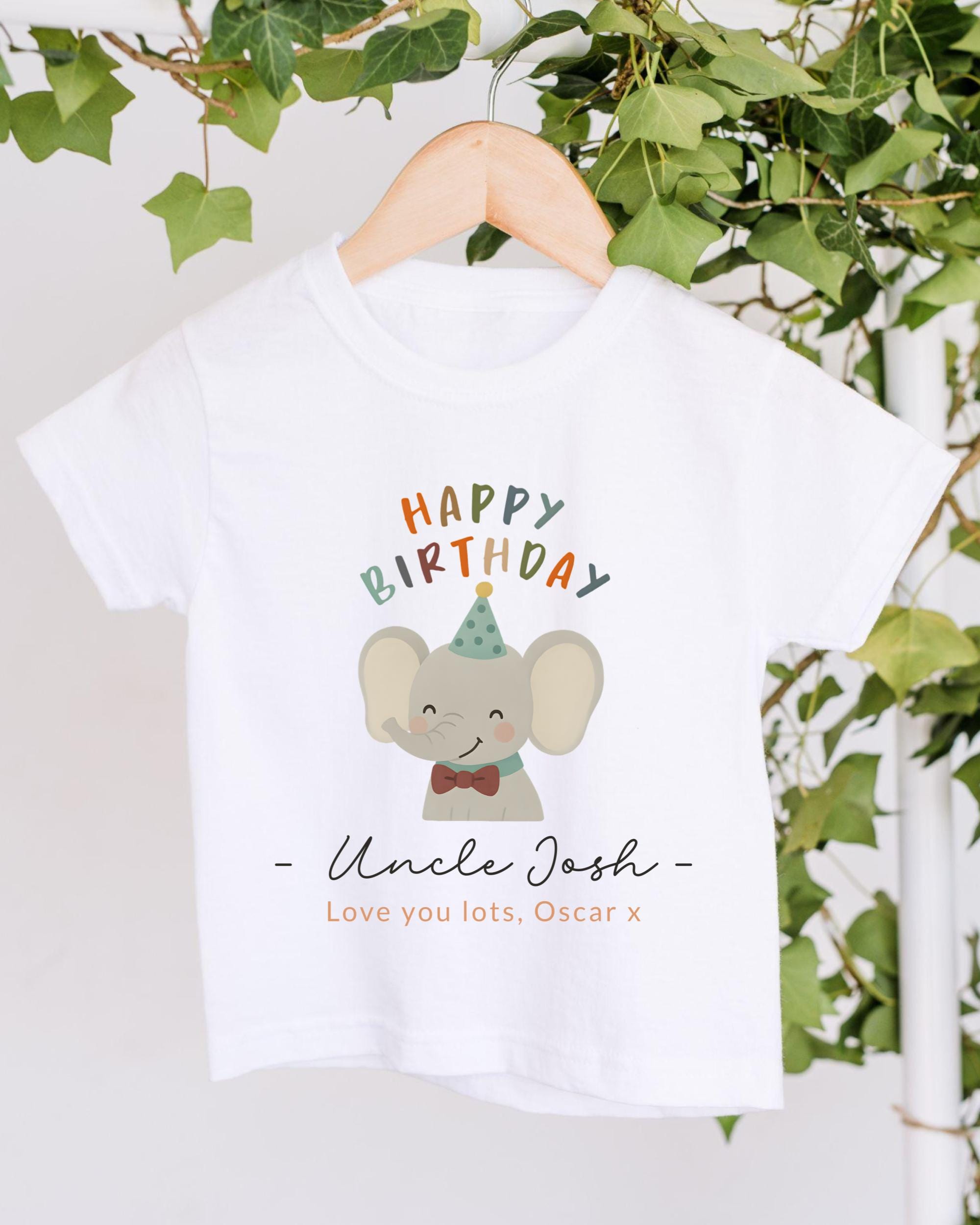 A white t-shirt with a colorful graphic of an elephant wearing a party hat and the text "Happy Birthday Uncle Josh" printed on it, hanging on a wooden hanger.