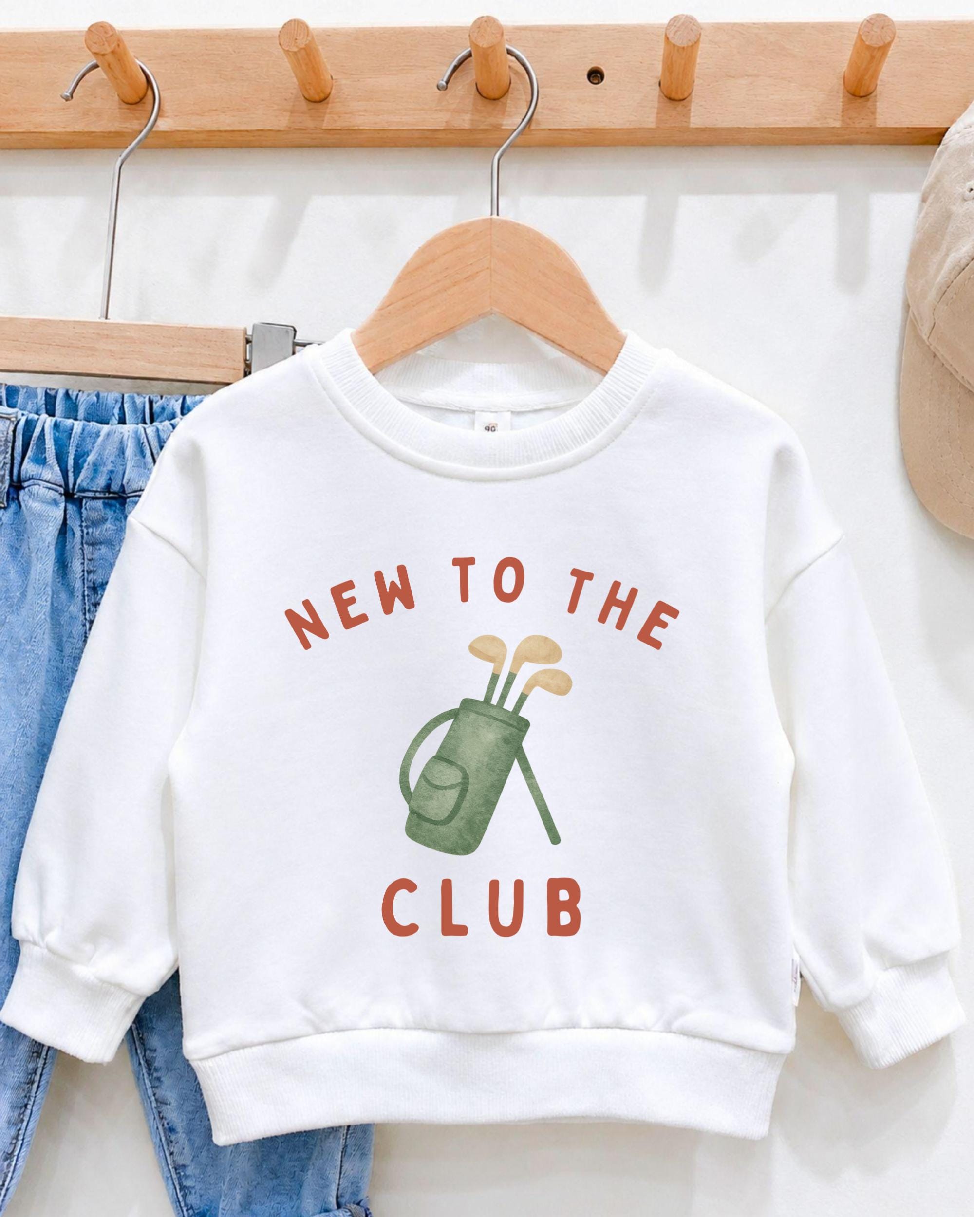 A white sweatshirt with the text "NEW TO THE CLUB" printed on it, along with an illustration of a golf club.