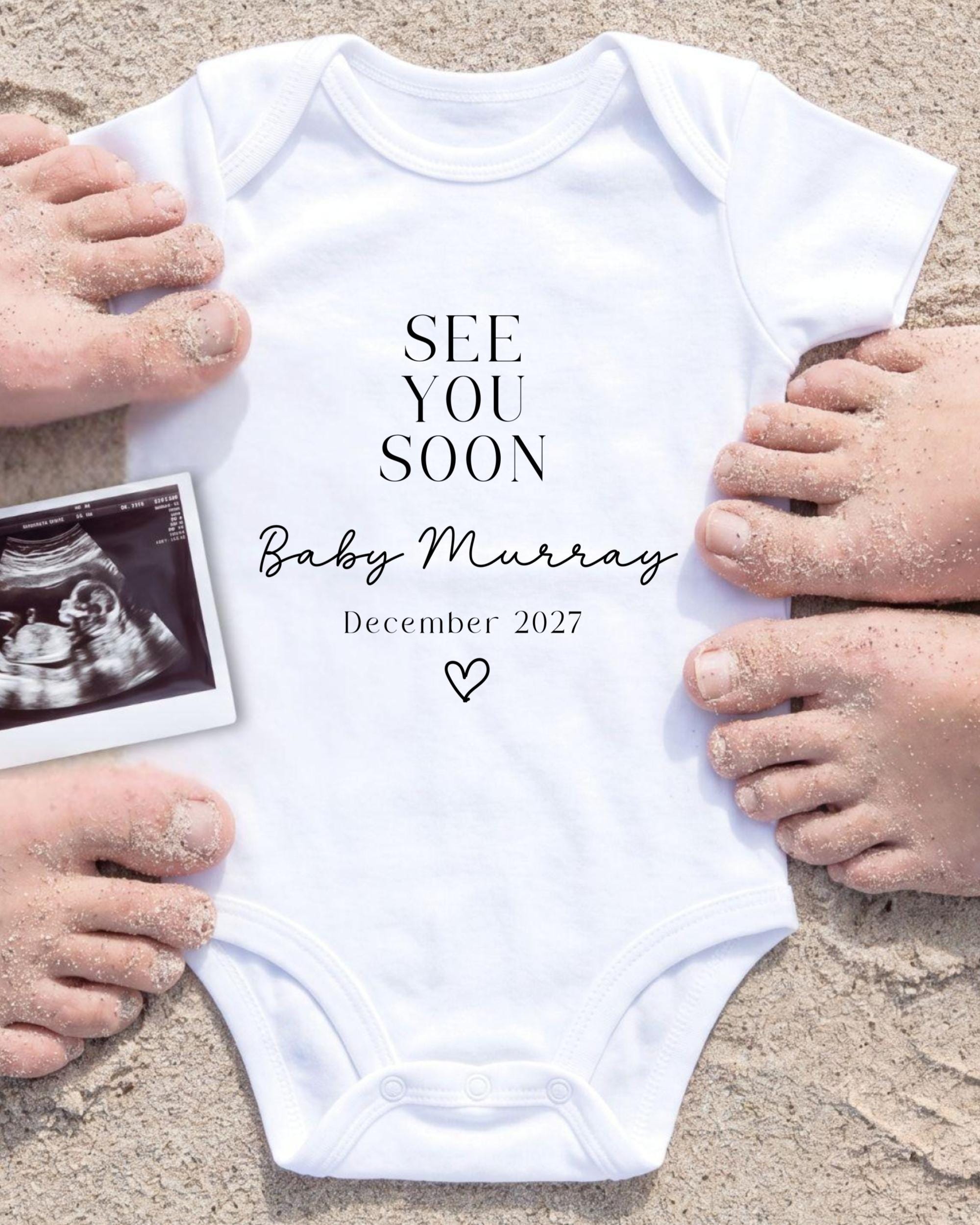 a white onesie with the text "See you soon Baby Murray" and the date "December 2027" printed on it, surrounded by sand and a black and white photograph of a baby's belly.