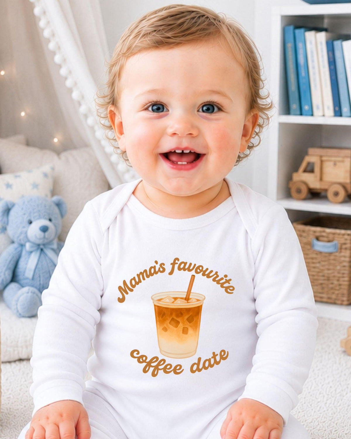A smiling baby wearing a white onesie with a coffee cup graphic and text, sitting in a room with a teddy bear and bookshelf in the background.