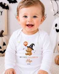 A smiling baby wearing a white onesie with a teddy bear and a witch hat, surrounded by Halloween decorations.
