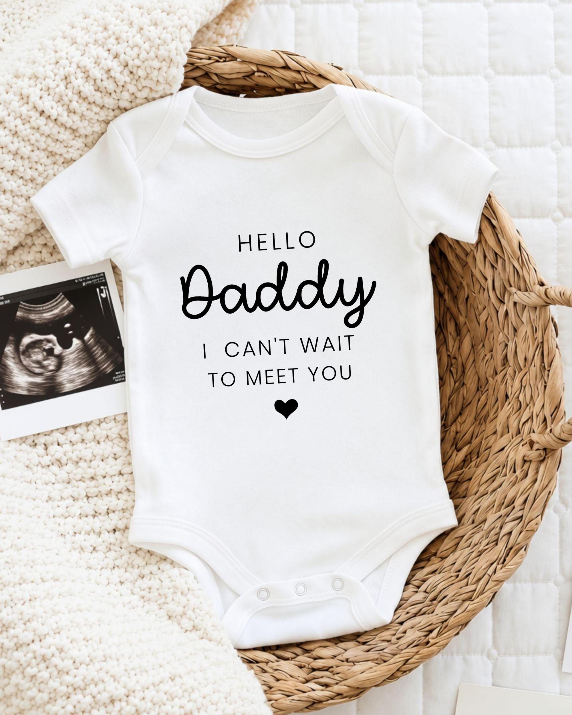 A white baby onesie with the text "Hello Daddy I can't wait to meet you" printed on it, placed on a woven basket.