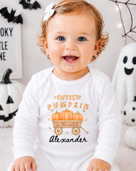 A smiling baby girl wearing a white onesie with a pumpkin design and text, sitting in front of Halloween decorations.