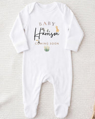 A white onesie with the text "BABY Harrison COMING SOON" printed on it, along with a small illustration of a giraffe and a plant.