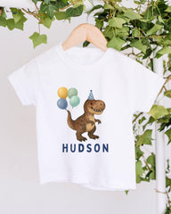 A white t-shirt with a cartoon dinosaur wearing a party hat and holding balloons, with the name "Hudson" printed below.