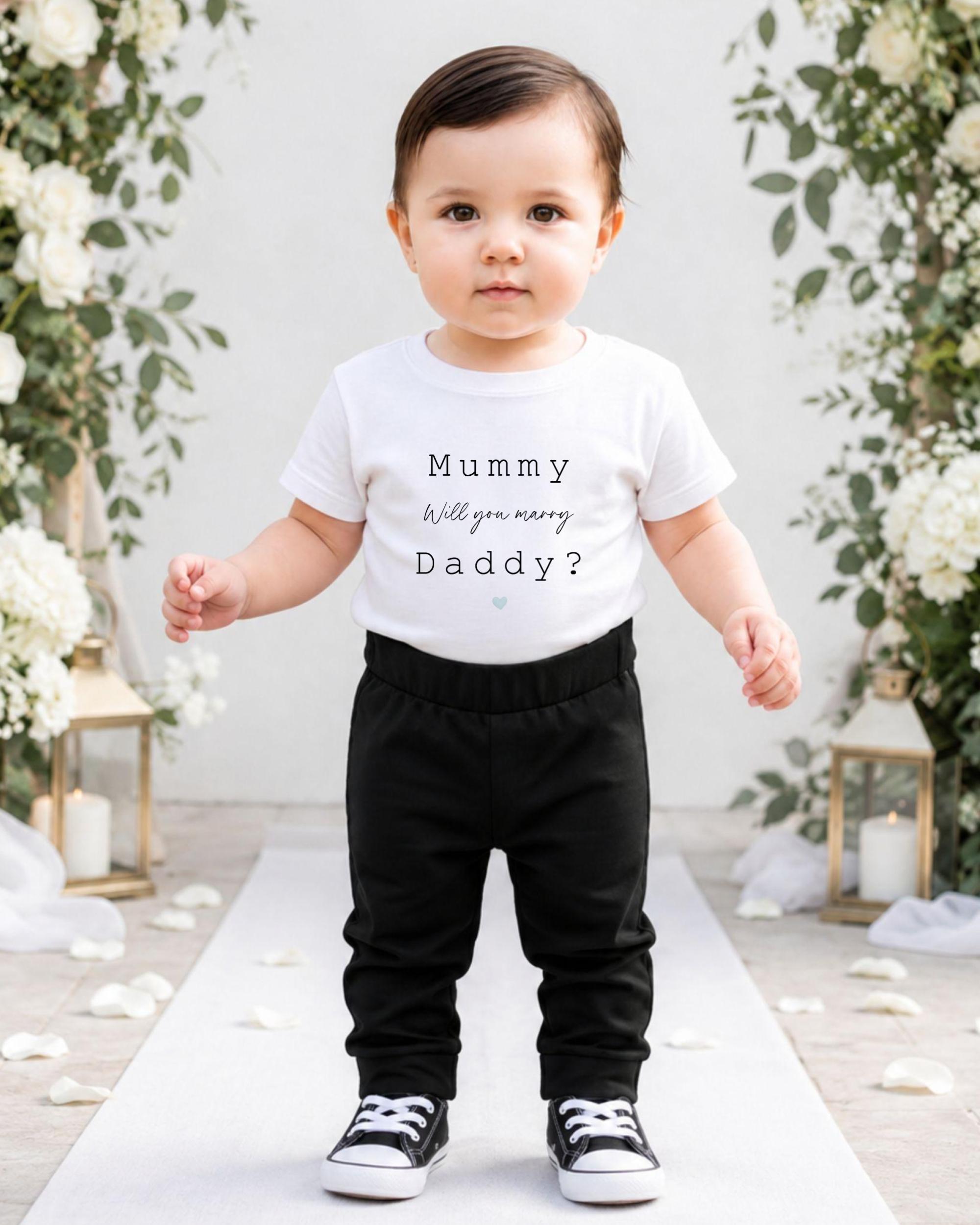 A young child wearing a white t-shirt with text and black pants stands in front of a floral archway.
