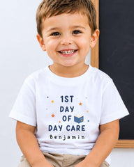 A young boy wearing a white t-shirt with the text "1st DAY OF DAY CARE Benjamin" printed on it, smiling at the camera.