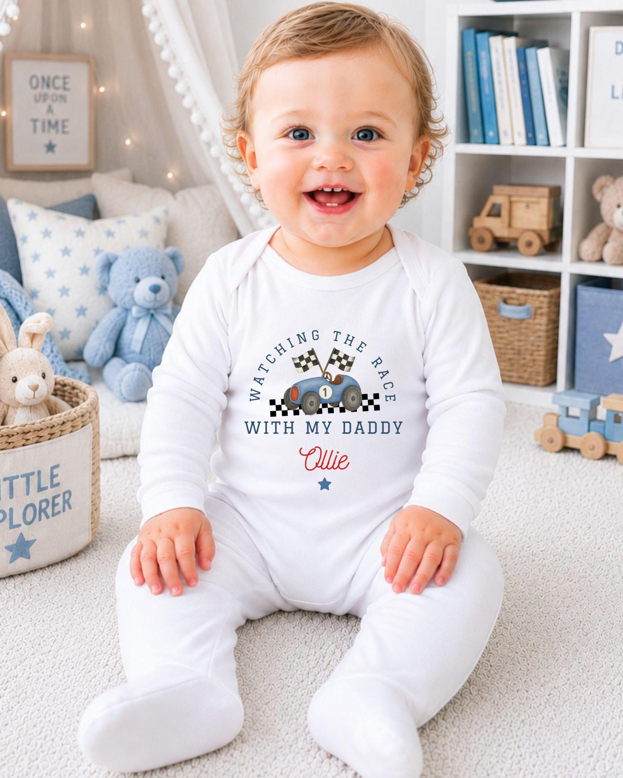 A smiling baby boy wearing a white onesie with a race car design and the text "Watching the race with my daddy" is sitting on the floor in a nursery setting.