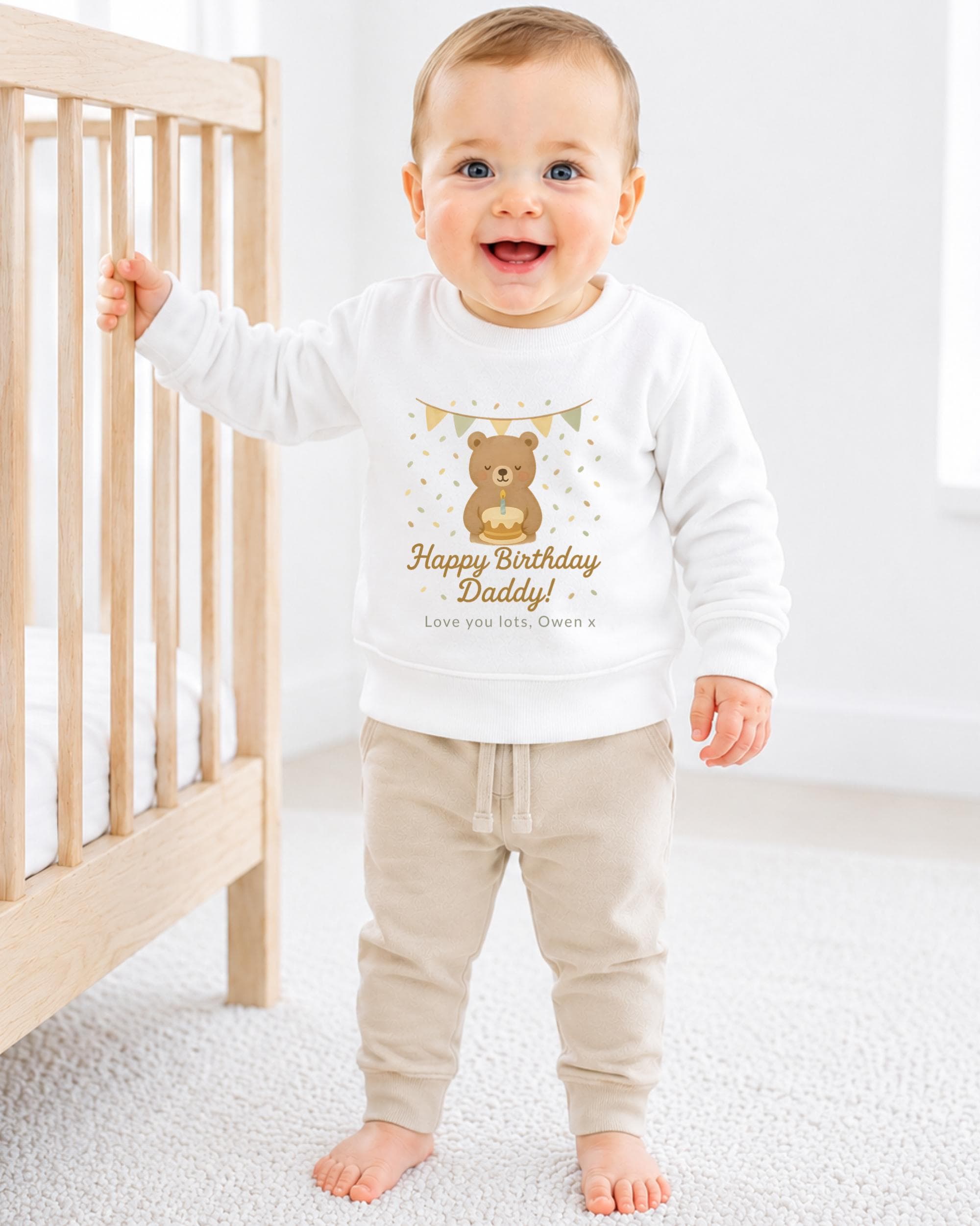 A smiling baby boy wearing a white sweatshirt with a teddy bear and birthday message, standing next to a wooden crib.