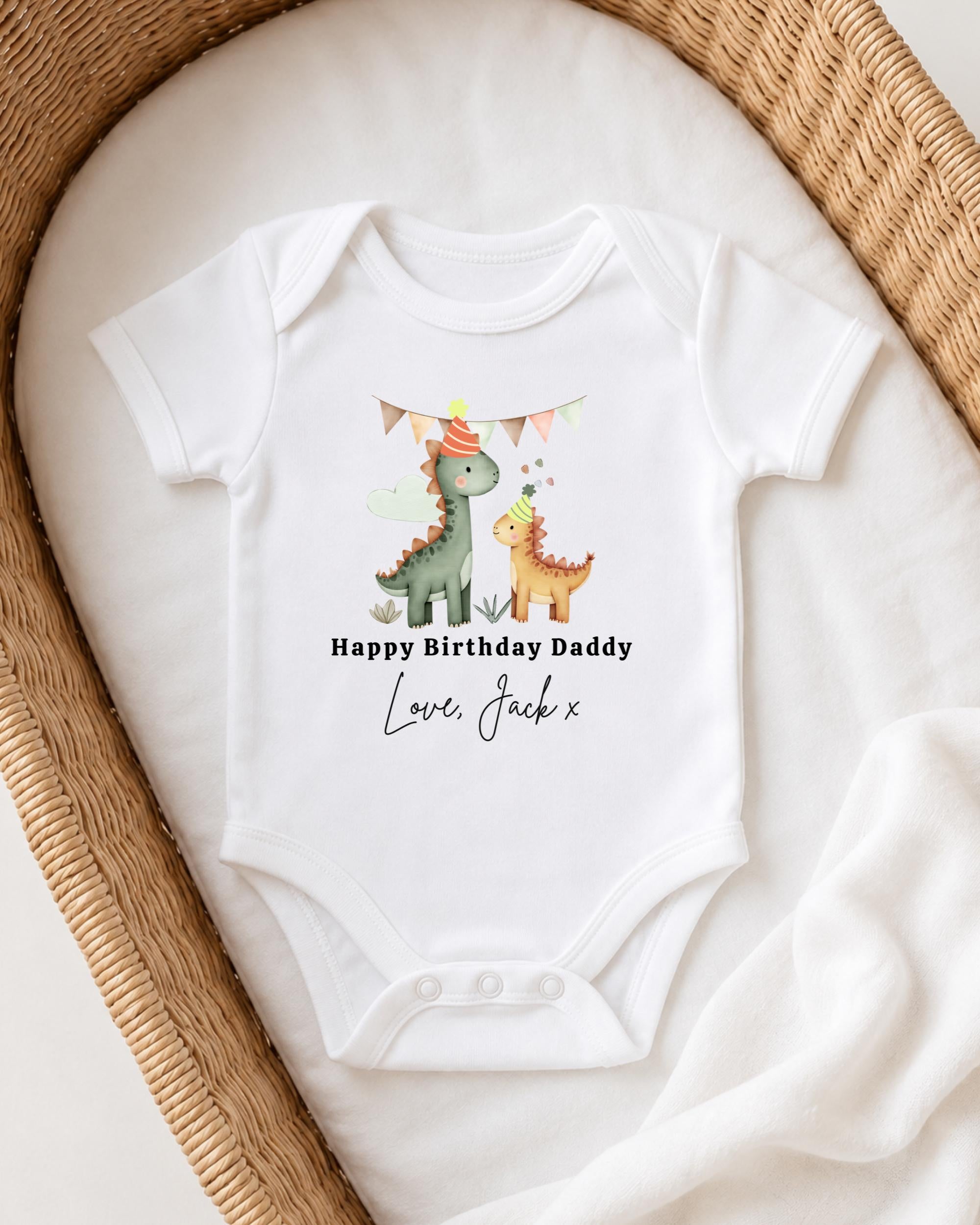A white onesie with a dinosaur design and text that reads "Happy Birthday Daddy Love Jack x" is displayed on a bed.